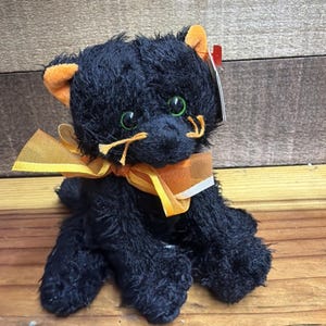 Peut inclure: Une peluche chat noire avec une fourrure texturée, des yeux vert vif et l'intérieur des oreilles orange. Le chat a des moustaches orange et porte un ruban orange transparent noué autour du cou. Une petite étiquette rouge et blanche est fixée à l'oreille gauche.