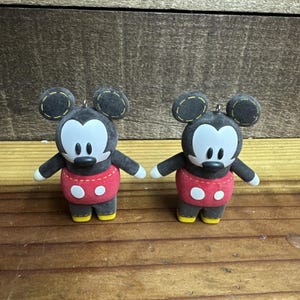 Puede incluir: Dos figuras de Mickey Mouse. Cada figura tiene un cuerpo negro, una cara blanca y pantalones cortos rojos con botones blancos. Las orejas son de color gris oscuro y los zapatos son amarillos. Las figuras están sobre una superficie de madera.