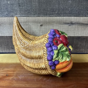 Puede incluir: Una cornucopia de cerámica llena de frutas y uvas coloridas. La cornucopia es marrón y tejida, asemejándose a una cesta.