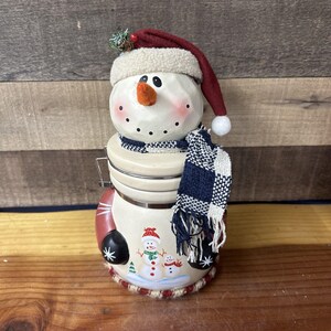 Pode incluir: Um pote de biscoitos de cerâmica em forma de boneco de neve com um chapéu de Papai Noel vermelho, um cachecol xadrez azul e branco e um desenho pintado de dois bonecos de neve menores na frente.