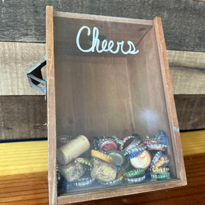 May include: A wooden box with a glass front, labeled "Cheers", filled with bottle caps and corks.