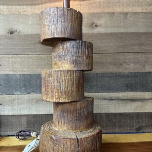 Op de afbeelding: Een rustieke houten lampvoet gemaakt van gestapelde boomstamschijven. De lampvoet is bruin en heeft een natuurlijke houtnerf. De lamp is aan en heeft een witte lampenkap.