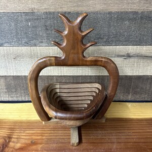 Puede incluir: Un frutero de madera con un diseño único y tallado. El frutero está hecho de madera oscura y tiene una serie de anillos concéntricos que crean un efecto escalonado. El frutero está soportado por una base que tiene la forma de un tronco de árbol estilizado.
