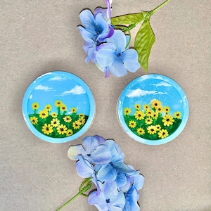 Puede incluir: Dos posavasos de cerámica azul y blanco con un diseño pintado de flores amarillas sobre fondo azul. Los posavasos están sobre una superficie marrón con flores azules.