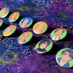 May include: A collection of oval-shaped character button pins with green borders. The pins feature various cartoon portraits of people with different hair colors and styles. Some pins are attached to gold star-shaped keychains. The background is a dark blue fabric with a celestial pattern.