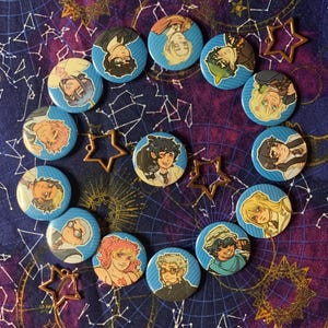 May include: A collection of blue button pins featuring various character illustrations, arranged in a circular pattern. Accompanying the pins are several gold-colored star-shaped charms. The background is a dark blue fabric with a celestial design.