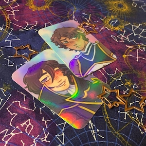 May include: Two iridescent holographic cards featuring anime-style illustrations of two characters with blushing cheeks. The cards are decorated with gold star charms.