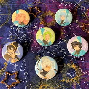 May include: A collection of six round pinback buttons featuring colorful anime-style character illustrations. Each button has a unique design with characters in hats and robes. The buttons are arranged on a dark blue fabric with a celestial pattern and star-shaped charms.
