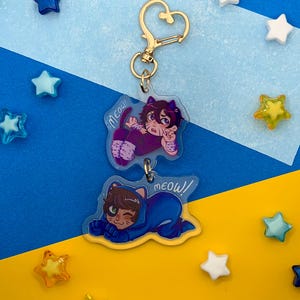 May include: A double-layered acrylic keychain featuring two cartoon characters in cat-themed outfits. The top character wears purple with cat ears, and the bottom character wears blue with cat ears. Both charms include the handwritten text Meow!
