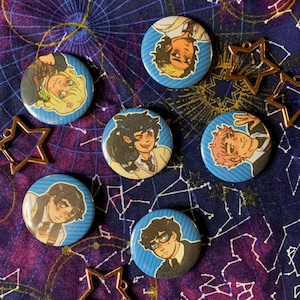 Puede incluir: Una colección de siete botones redondos azules con ilustraciones de personajes de estilo anime. Los botones tienen un fondo azul con líneas diagonales blancas. Los botones están dispuestos sobre una tela azul oscuro con un patrón celestial, junto con dijes dorados en forma de estrella.