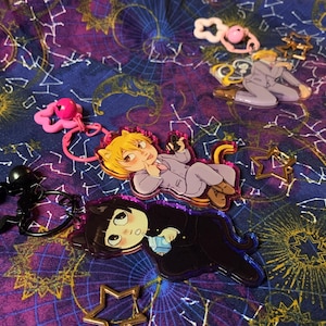 May include: A selection of anime-style keyrings with character designs in different poses. The keyrings have a glossy finish and are shaped like stars, cats, and other forms. The background is a dark blue fabric with a celestial pattern.