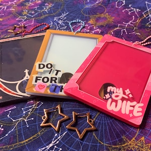 Op de afbeelding: Drie rechthoekige badgehouders in zwart, oranje en roze, elk met een helder venster. De oranje houder heeft de tekst "DO IT FOR" en een hart. De roze houder zegt "my WIFE". Twee bronzen sterbedels op de voorgrond.