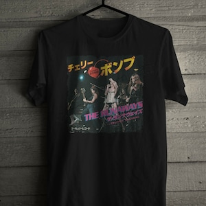 Può includere: Maglietta nera con una grafica di una band chiamata "The Runaways" con il testo "Cherry Bomb" e "The Runaways" in giapponese. I membri della band sono ritratti sul palco con microfoni e chitarre.