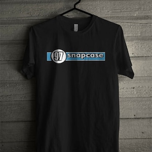 May include: Black t-shirt with the text "snapcase" in white letters on a light blue background. The number "97" is inside a white circle on the left side of the text. The t-shirt is hanging on a black hanger against a wooden background.