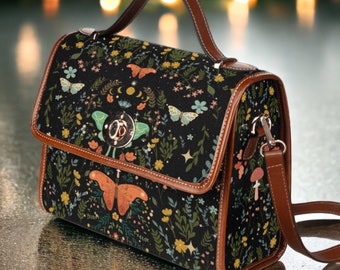 Könnte beinhalten: Eine schwarze Satchel-Tasche mit braunem Lederbesatz und einem floralen Druck mit Schmetterlingen, Monden und Sternen. Die Tasche hat einen oberen Griff und einen langen, verstellbaren Schultergurt.