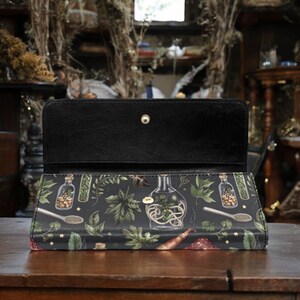 May include: A black leather wallet with a zipper closure and a botanical print featuring illustrations of glass bottles filled with herbs and flowers. The print is on a dark green background with gold stars.
