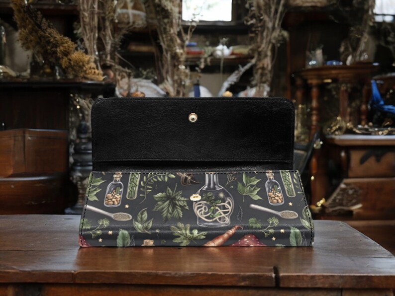May include: A black leather wallet with a zipper closure and a botanical print featuring illustrations of glass bottles filled with herbs and flowers. The print is on a dark green background with gold stars.