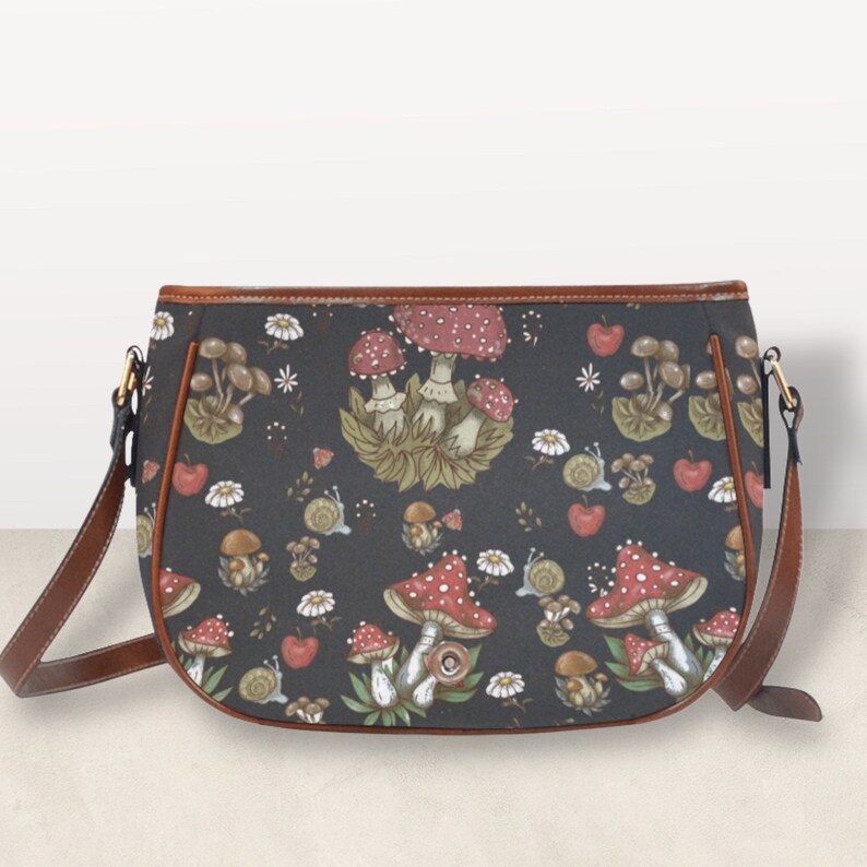 May include: A black leather shoulder bag with a brown trim and a mushroom print. The print features red and white mushrooms with green leaves and stems.