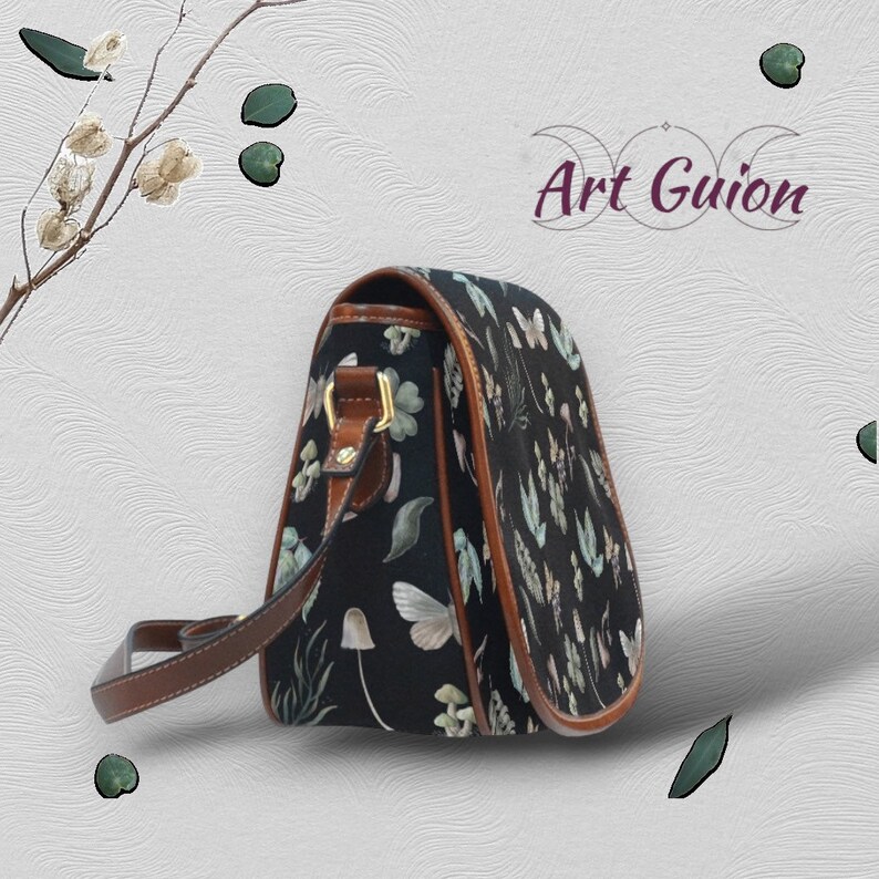 Puede incluir: Un bolso bandolera negro con una correa y solapa de cuero marr&oacute;n. El bolso tiene un estampado floral con mariposas, setas y hojas.