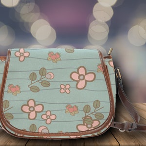 May include: A light blue crossbody bag with a floral pattern featuring pink flowers and green leaves. The bag has brown leather trim and a matching adjustable strap. The bag is approximately 10 inches wide.