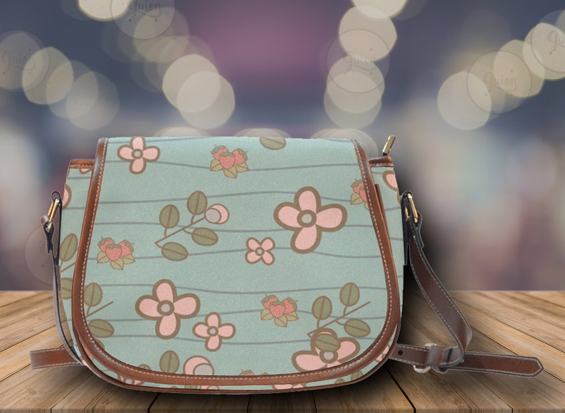 May include: A light blue crossbody bag with a floral pattern featuring pink flowers and green leaves. The bag has brown leather trim and a matching adjustable strap. The bag is approximately 10 inches wide.