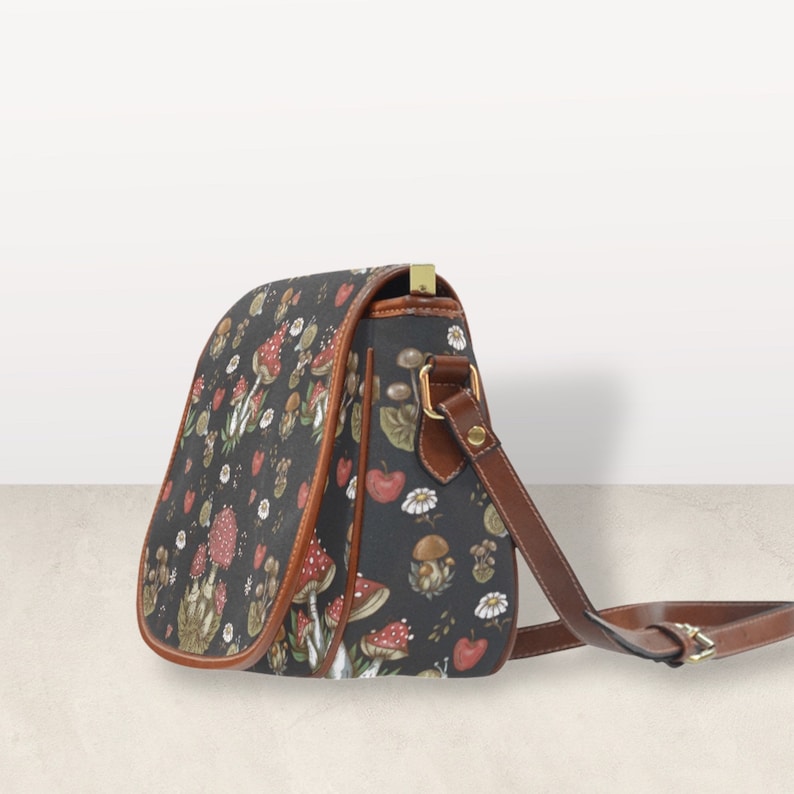 May include: A black leather shoulder bag with a brown strap. The bag is decorated with a pattern of mushrooms, leaves, and flowers.