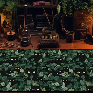 May include: A rectangular desk mat featuring a repeating pattern of black cat faces peeking out from green leaves on a black background. The mat is placed on a wooden desk with candles and other decorative items.