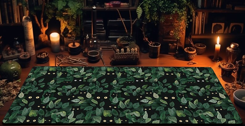 May include: A rectangular desk mat featuring a repeating pattern of black cat faces peeking out from green leaves on a black background. The mat is placed on a wooden desk with candles and other decorative items.