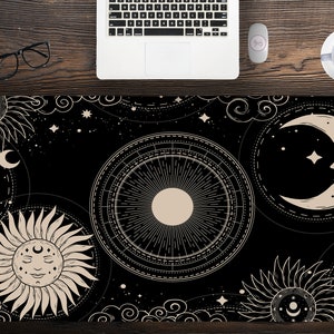 May include: Black desk mat with a celestial design featuring a sun, moon, and stars. The mat has a detailed, cream-colored illustration on a black background. A laptop, mouse, glasses, and a cup of coffee are on a wooden desk.