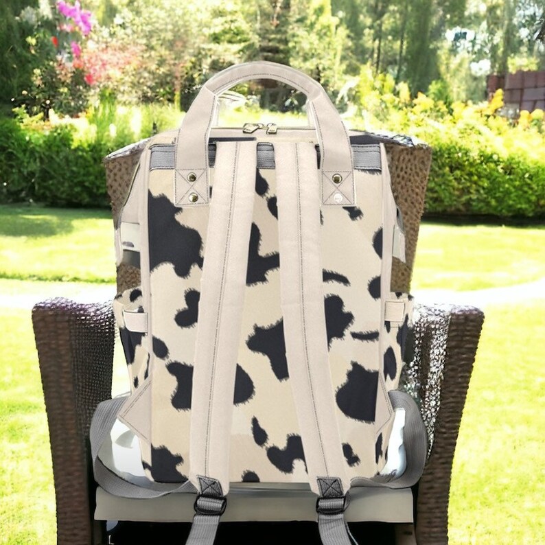 K&ouml;nnte beinhalten: Ein wei&szlig;er und schwarzer Rucksack aus Canvas mit Kuhmuster und verstellbaren Tr&auml;gern. Der Rucksack hat einen Rei&szlig;verschluss und mehrere Taschen.