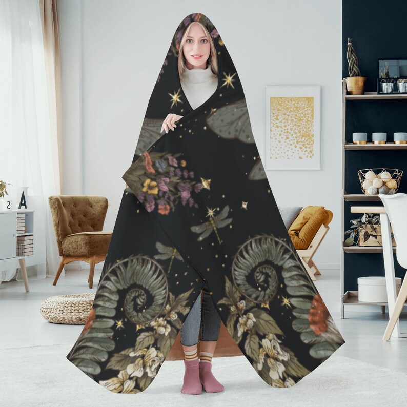 May include: A hooded blanket with a dark background featuring a botanical and celestial design. The pattern includes flowers, dragonflies, and stars in shades of green, purple, and gold. The blanket is worn by a person.