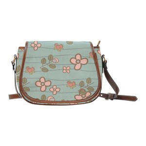 May include: A floral-patterned crossbody bag with a light blue background and brown trim. The bag features a repeating pattern of pink flowers and green leaves. The adjustable strap and flap closure are brown.