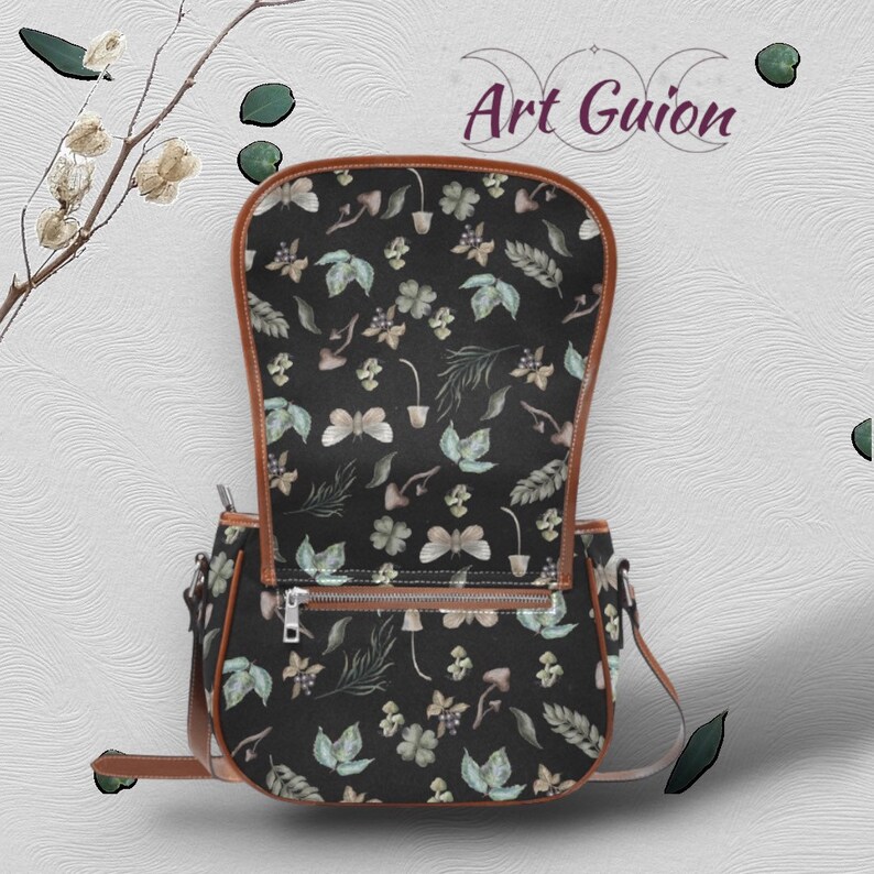 Puede incluir: Un bolso bandolera de cuero marr&oacute;n con cierre de solapa. El bolso tiene un forro de tela negro con un estampado floral con mariposas, hojas y setas.