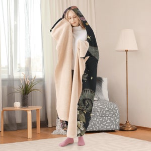 May include: A hooded blanket with a black exterior featuring floral and celestial designs, and a cream-colored sherpa interior. The blanket has a hood and is worn by a person.