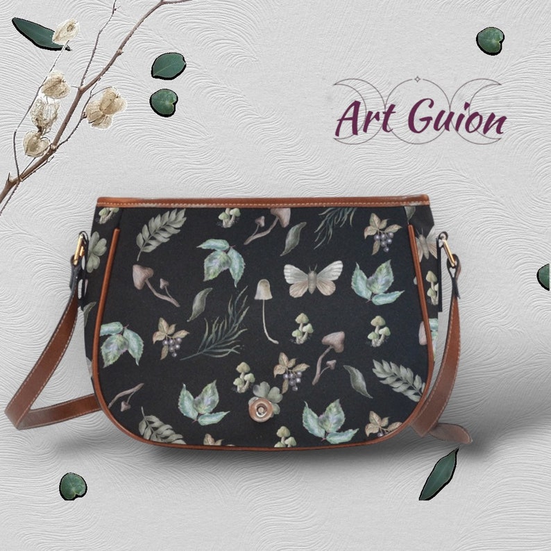 Puede incluir: Un bolso de hombro negro con una correa marr&oacute;n y un estampado floral con mariposas, setas y hojas. El bolso tiene una forma curva y un cierre de solapa.