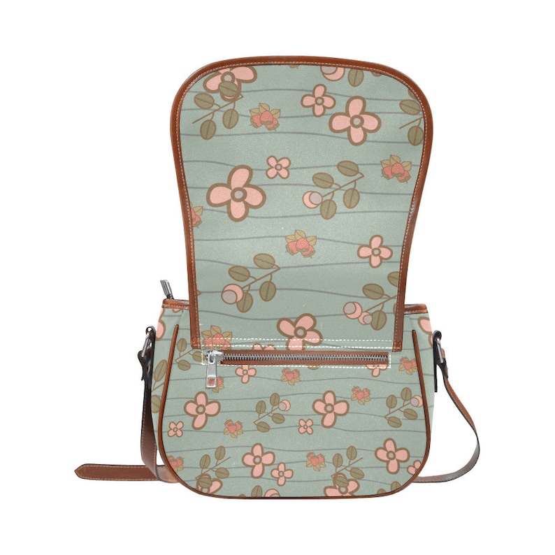 May include: A floral-patterned crossbody bag with a light green background and brown trim. The bag features a repeating pattern of pink flowers and brown stems. The interior is visible, and the bag has a zipper closure and a brown strap.