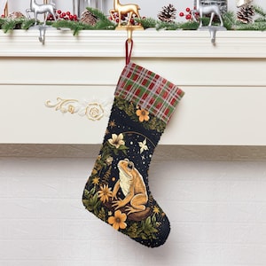 May include: A Christmas stocking with a dark blue background, featuring a detailed illustration of a frog surrounded by flowers. The top of the stocking has a red and green plaid pattern. The stocking is hanging from a white mantelpiece.