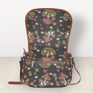 May include: A brown leather shoulder bag with a black fabric flap featuring a repeating pattern of red and white mushrooms with green stems and leaves. The bag has a zipper closure and a brown adjustable strap.