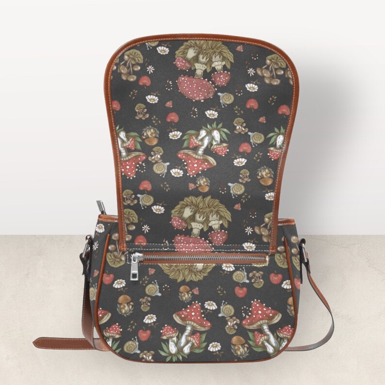 May include: A brown leather shoulder bag with a black fabric flap featuring a repeating pattern of red and white mushrooms with green stems and leaves. The bag has a zipper closure and a brown adjustable strap.