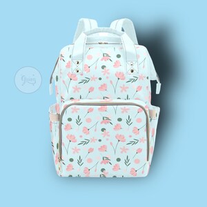 以下が含まれることがあります： ピンクの花と緑の葉が描かれた花柄の薄い青色のオムツバッグ。バッグには金色のジッパーと金色のアクセントが付いています。