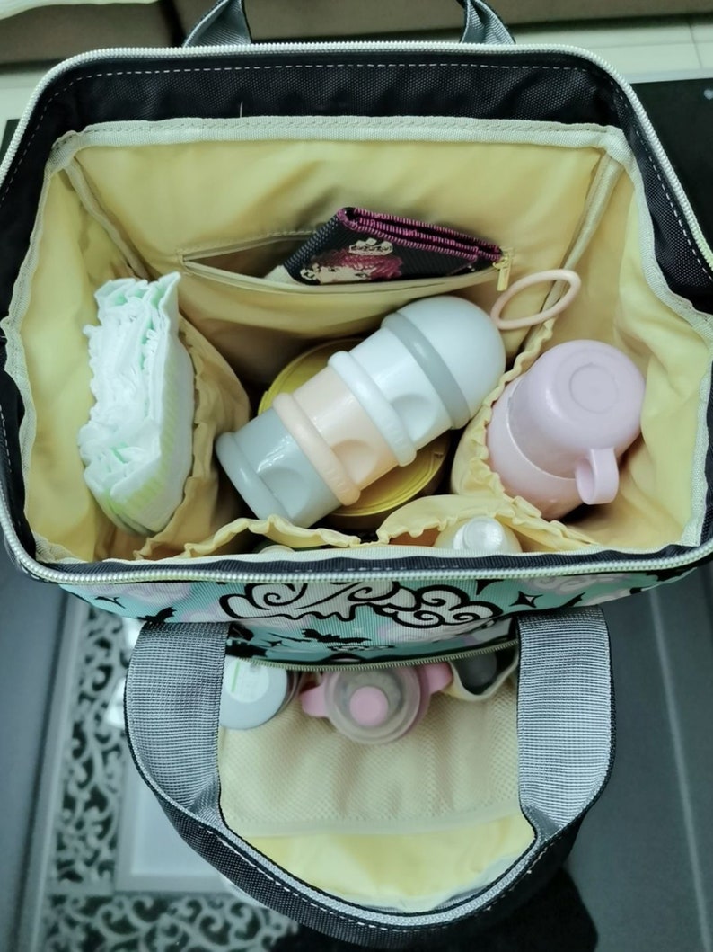 K&ouml;nnte beinhalten: Eine schwarze Wickeltasche mit Blumendruck und gelber Innenausstattung. Die Tasche ist offen und mit verschiedenen Babyartikeln gef&uuml;llt, darunter eine Windel, eine Flasche, ein kleines Beh&auml;ltnis und eine rosa Tasche.