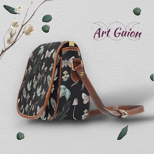 Puede incluir: Un bolso bandolera de lona negra con una correa y solapa de cuero marr&oacute;n. El bolso presenta un estampado floral y de mariposas. El bolso est&aacute; sobre un fondo blanco texturizado.