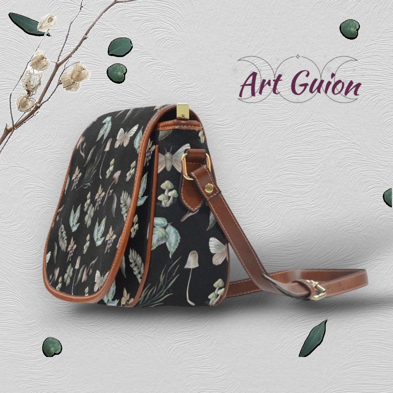 Puede incluir: Un bolso bandolera de lona negra con una correa y solapa de cuero marr&oacute;n. El bolso presenta un estampado floral y de mariposas. El bolso est&aacute; sobre un fondo blanco texturizado.