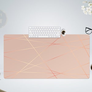Puede incluir: Una alfombrilla de escritorio rosa claro con un patrón geométrico de finas líneas doradas. La alfombrilla está sobre una superficie blanca con un teclado blanco, un ratón blanco, unas gafas negras y una taza de café.