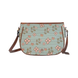 May include: A light blue crossbody bag with a floral pattern. The bag features pink flowers and brown stems against a light blue background with horizontal lines. It has a brown leather strap and trim.