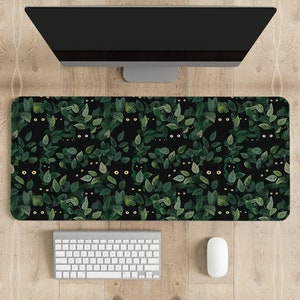 May include: A rectangular desk mat featuring a repeating pattern of black cats peeking out from green leaves on a black background. A white keyboard and mouse are below a computer monitor.