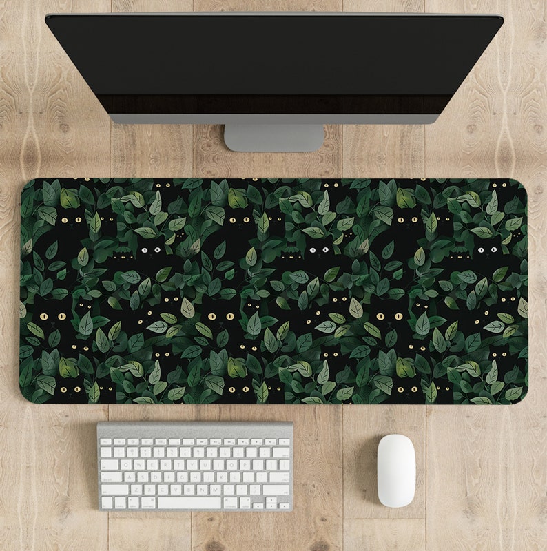 May include: A rectangular desk mat featuring a repeating pattern of black cats peeking out from green leaves on a black background. A white keyboard and mouse are below a computer monitor.