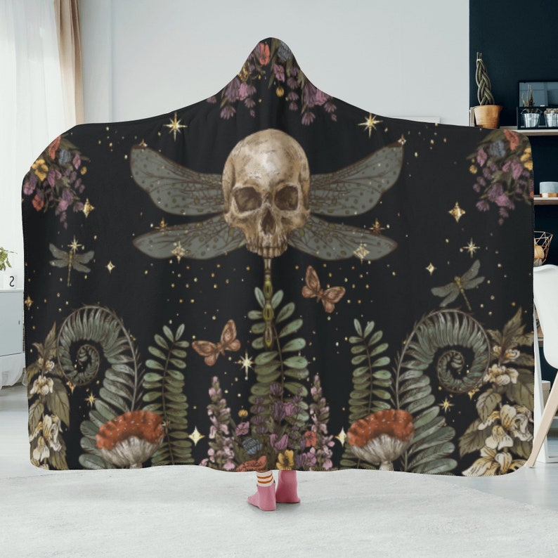 May include: Black hooded blanket featuring a skull with dragonfly wings, butterflies, and botanical elements. The design includes ferns, flowers, and stars. The blanket is made of soft material and is suitable for snuggling.