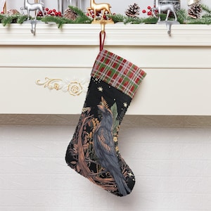 May include: Black Christmas stocking with a raven illustration. The stocking has a plaid cuff in red, green, and brown. The raven is perched on a tree branch with a forest background. The stocking is hanging from a white mantel.
