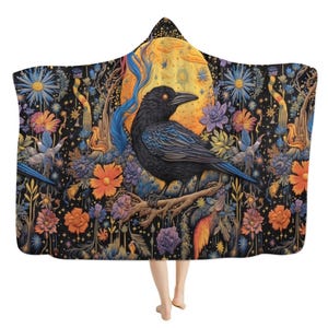 May include: A hooded cloak with a detailed illustration of a black raven perched on a branch, set against a full moon and colourful flowers. The cloak has a black background with vibrant floral and celestial elements.